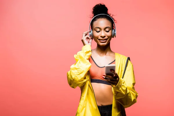 Bela sorridente afro-americana esportista em fones de ouvido usando smartphone isolado em coral com espaço de cópia — Fotografia de Stock