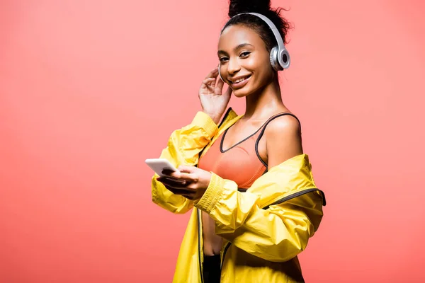 Beautiful smiling african american sportswoman in headphones looking at camera and using smartphone isolated on coral — Stock Photo