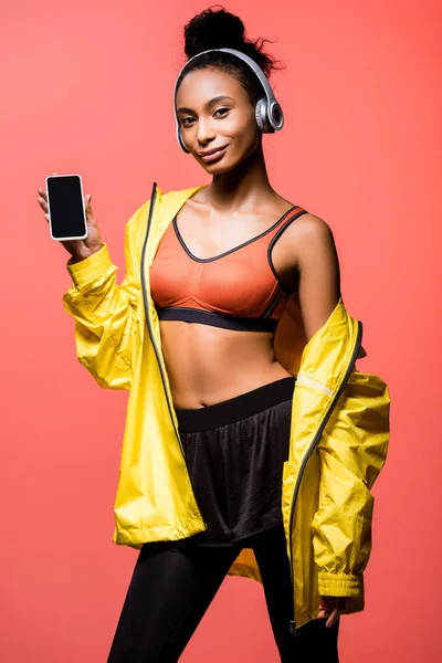 Hermosa deportista afroamericana en auriculares mirando a la cámara y presentando teléfono inteligente con pantalla en blanco aislado en coral - foto de stock