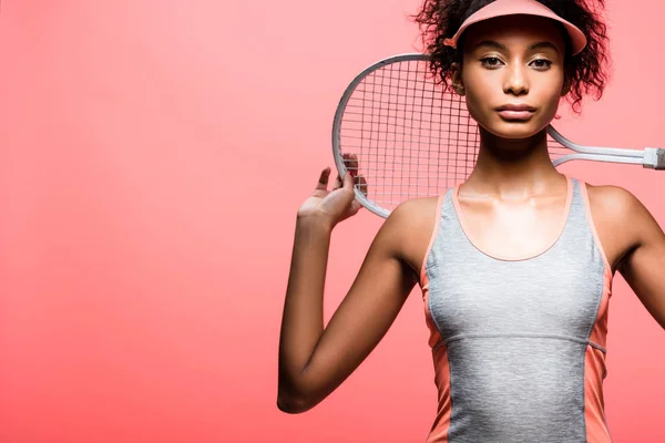 African american sportswoman in sun visor holding tennis racket and looking at camera isolated on coral with copy space — Stock Photo