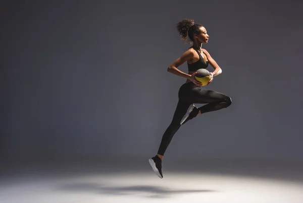 Vue latérale d'une sportive afro-américaine sportive tenant le ballon et sautant sur le gris — Photo de stock