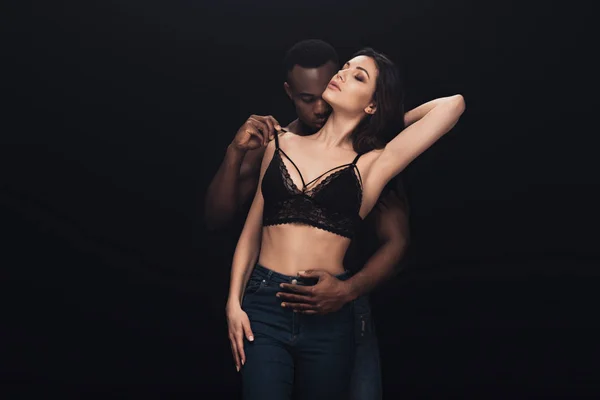 African american man passionately hugging sexy woman in lace bra isolated on black with copy space — Stock Photo
