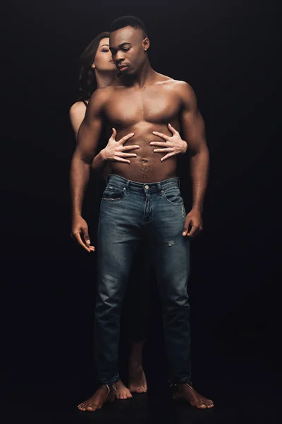 Hermosa sexy mujer abrazando sin camisa africano americano hombre en denim aislado en negro - foto de stock