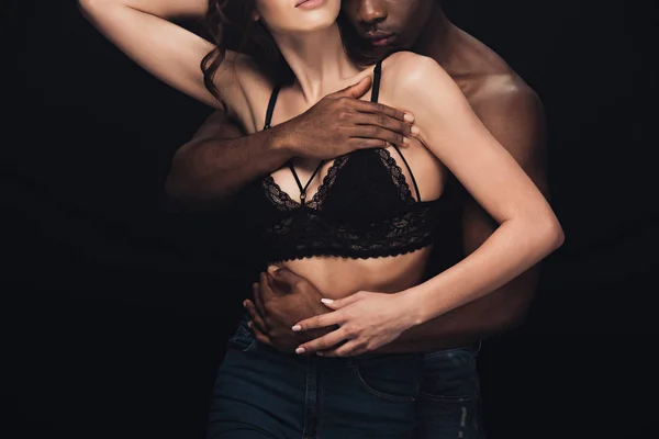 Recortado vista de africano americano hombre abrazando sexy mujer en lencería aislado en negro - foto de stock