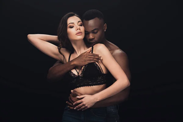 Africano americano hombre apasionadamente abrazando sexy mujer en encaje sujetador aislado en negro - foto de stock