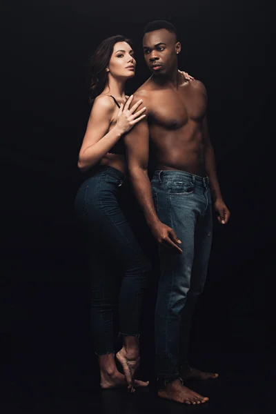 Hermosa mujer seductora abrazando sin camisa hombre afroamericano en denim aislado en negro - foto de stock