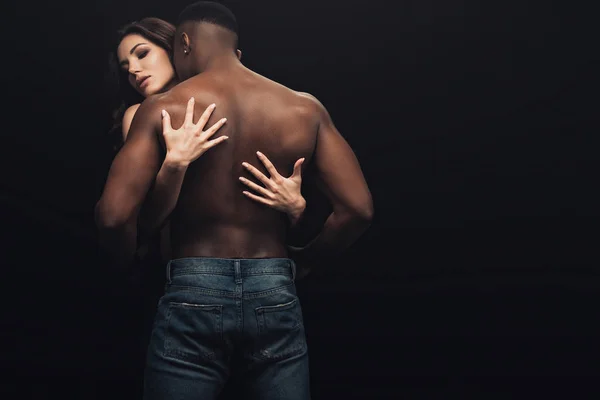 Vista posteriore di bella donna sexy abbracciando uomo afroamericano senza maglietta isolato su nero — Foto stock