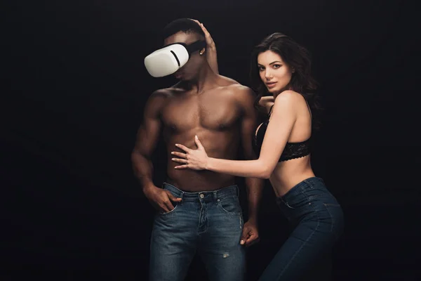 Beautiful sexy woman posing and looking at camera near african american man in virtual reality headset isolated on black — Stock Photo