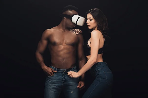 Красивая сексуальная женщина, позирующая рядом с африканским мужчиной без рубашки в наушниках виртуальной реальности, изолированных на черном — стоковое фото