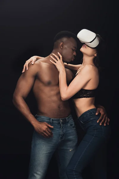 Без сорочки афроамериканський чоловік обіймає сексуальну жінку в гарнітурі віртуальної реальності ізольовано на чорному — стокове фото