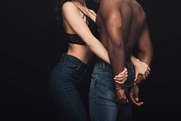 Частковий вид сексуальної жінки, що обіймає без сорочки афроамериканця в наручниках ізольовано на чорному — стокове фото