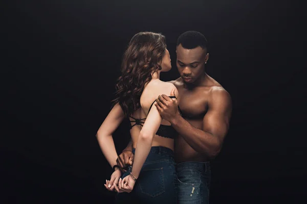 Hemdlose afrikanisch-amerikanische Mann umarmt schöne sexy Frau in Handschellen isoliert auf schwarz mit Kopierraum — Stockfoto