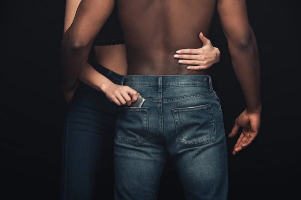 Vue recadrée de la femme embrassant torse nu homme afro-américain et tenant préservatif isolé sur noir — Photo de stock