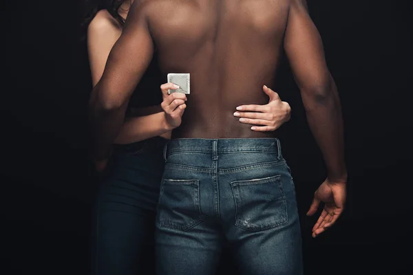 Visión parcial de la mujer abrazando al hombre afroamericano sin camisa y sosteniendo el condón aislado en negro - foto de stock