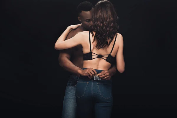 Afro-américain homme embrassant femme sexy en lingerie et tenant préservatif isolé sur noir — Photo de stock