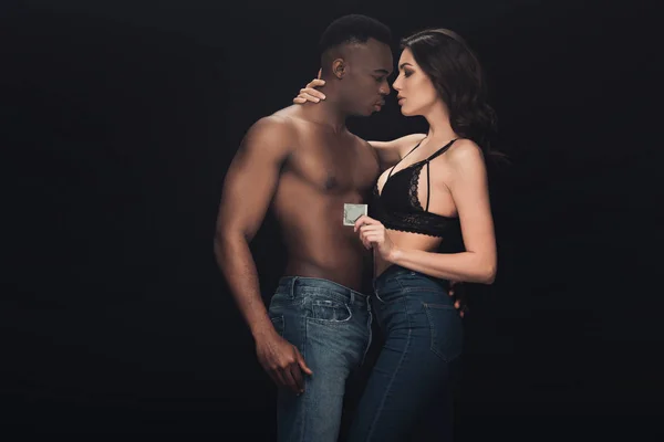 Bela mulher sexy abraçando sem camisa afro-americano homem e segurando preservativo isolado no preto — Fotografia de Stock