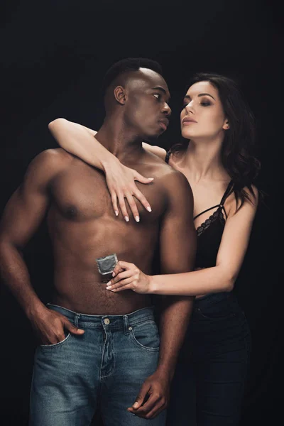 Sensual mulher abraçando sem camisa afro-americano homem e segurando preservativo isolado no preto — Fotografia de Stock