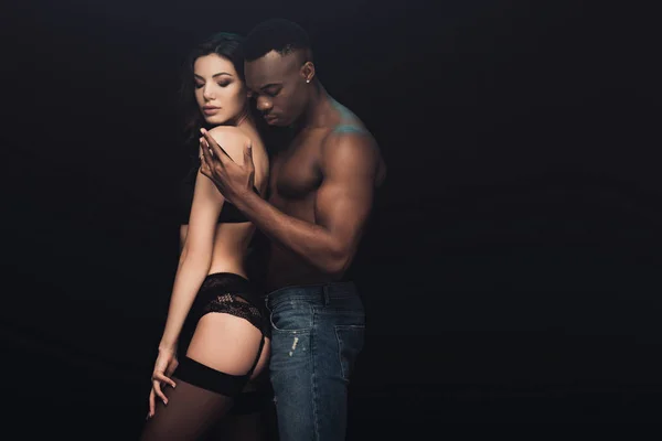 Africano americano hombre abrazando hermosa sexy mujer en lencería aislado en negro con copia espacio - foto de stock