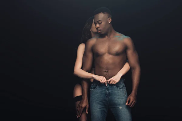 Сексуальная женщина обнимает и раздевает африканского мужчину без рубашки, изолированного от черного — стоковое фото
