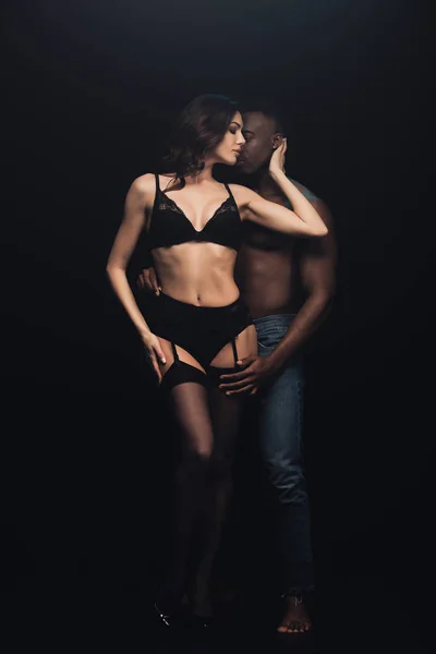 Africano americano hombre abrazando hermosa sexy mujer en lencería aislado en negro - foto de stock