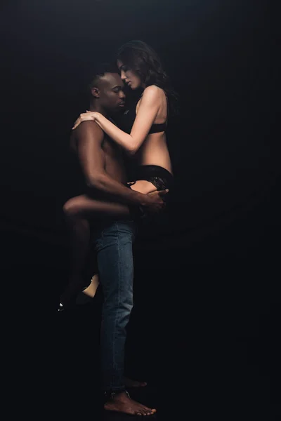 Hombre afroamericano sosteniendo en brazos hermosa mujer sexy en lencería aislada en negro con espacio de copia - foto de stock