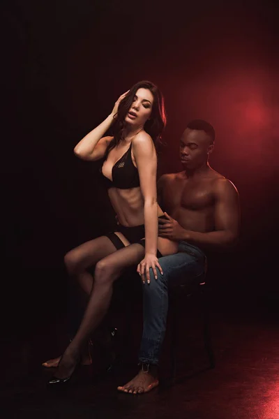 Beautiful sexy woman in lingerie sitting on shirtless african american man on black with red light — Stock Photo