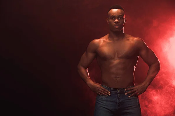 Sexy muscular afroamericano hombre en denim mirando a la cámara en la oscuridad con luz roja y espacio de copia - foto de stock