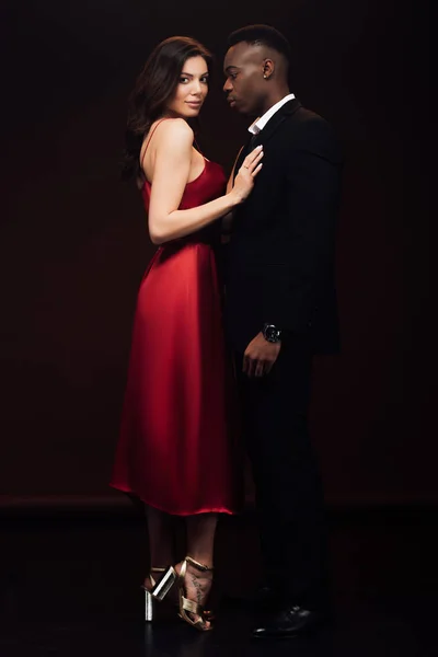 Belle femme en robe rouge à la mode posant avec l'homme afro-américain en costume isolé sur noir — Photo de stock