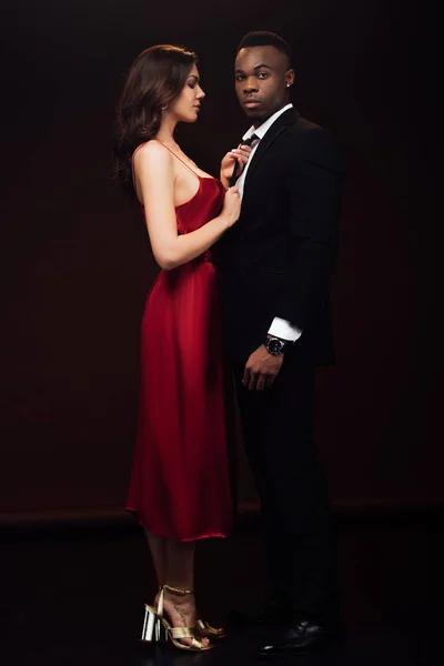 Hermosa mujer en vestido rojo posando con hombre afroamericano en traje aislado en negro - foto de stock