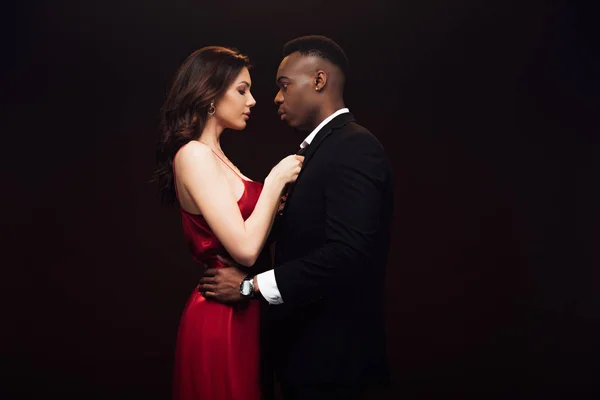 Hermosa mujer en vestido rojo posando con hombre afroamericano en traje aislado en negro con espacio de copia - foto de stock