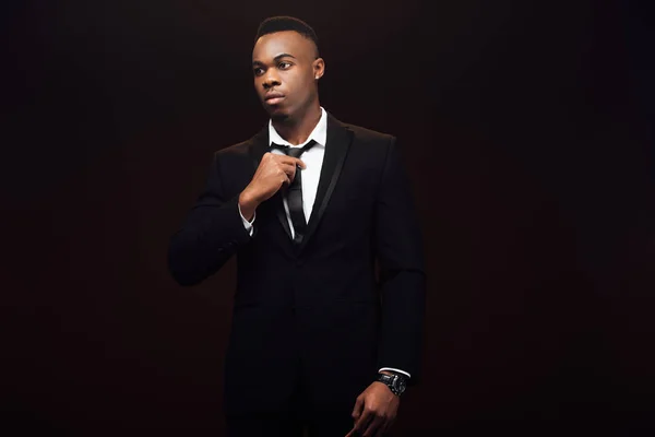 Handsome fashionable african american man in suit adjusting tie isolated on black — Stock Photo