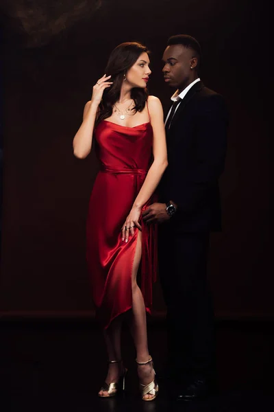 Hermosa pareja interracial en formal desgaste mirando el uno al otro y posando aislado en negro - foto de stock
