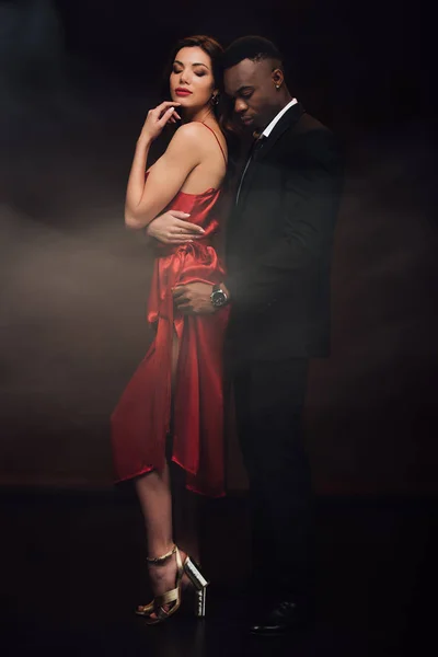 Afroamericano uomo abbracciando bella donna in abito rosso su nero con fumo — Foto stock