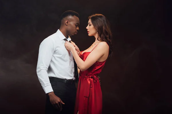 Beautiful woman in red dress undressing shirt of african american man isolated on black with copy space — Stock Photo