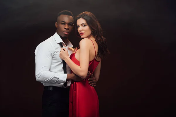 Красива жінка в червоній сукні дивиться на камеру і роздягається сорочка афроамериканця ізольована на чорному — стокове фото