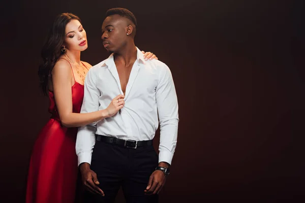 Beautiful woman in red dress undressing african american man isolated on black with copy space — Stock Photo