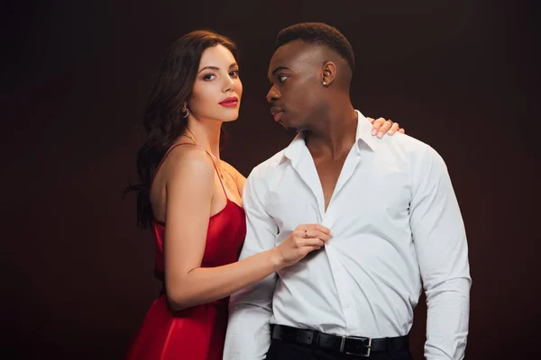 Bela mulher em vestido vermelho despir afro-americano homem isolado em preto — Fotografia de Stock