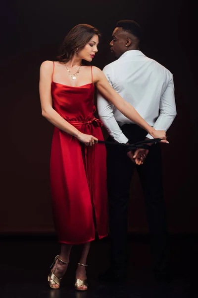 Beautiful woman in red dress tying hands of african american man with ribbon isolated on black — Stock Photo