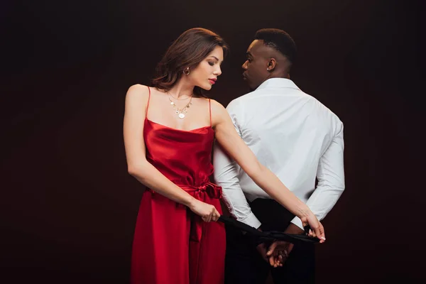 Beautiful woman in red dress tying hands of african american man with ribbon isolated on black — Stock Photo