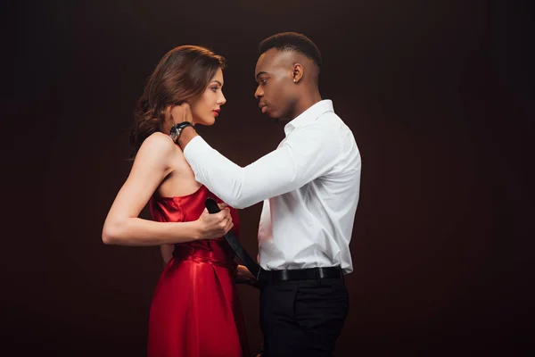 Hermosa pareja interracial en formal desgaste abrazando y mirando el uno al otro aislado en negro con espacio de copia - foto de stock