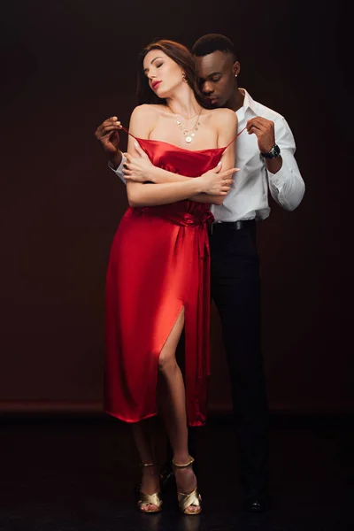 African american man undressing beautiful woman in red dress isolated on black — Stock Photo
