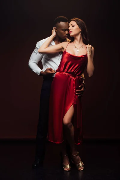 Bonito afro-americano homem abraçando bela mulher em vestido vermelho isolado no preto — Fotografia de Stock