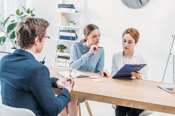 Attractive recruiters looking at clipboard near man sitting with clenched hands — Stock Photo