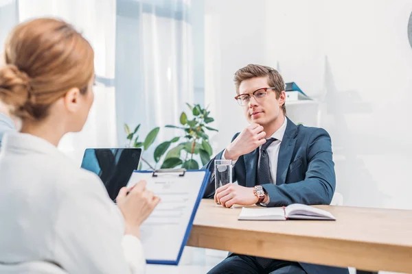Hombre pensativo en gafas sentado cerca del reclutador con portapapeles en la entrevista de trabajo - foto de stock