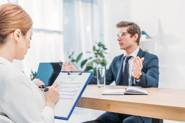 Selektiver Fokus des Recruiters, der auf Klemmbrett schreibt, in der Nähe eines Mannes, der im Vorstellungsgespräch spricht — Stockfoto