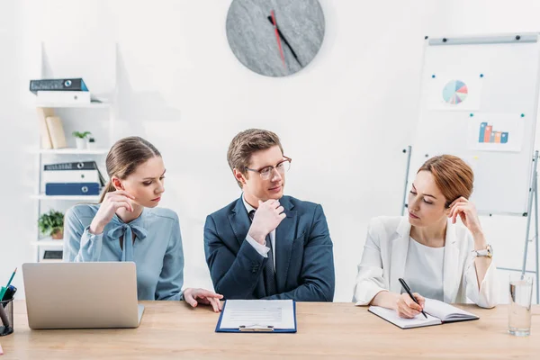 Pensive recruiter in glasses looking at coworker holding pen near notebook — Stock Photo