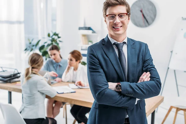 Enfoque selectivo de reclutador alegre en gafas de pie con brazos cruzados cerca de compañeros de trabajo - foto de stock