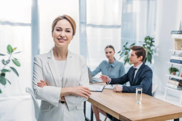 Enfoque selectivo de la mujer alegre de pie con brazos cruzados cerca de compañeros de trabajo - foto de stock