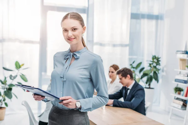 Enfoque selectivo de la mujer alegre de pie con portapapeles cerca de compañeros de trabajo - foto de stock