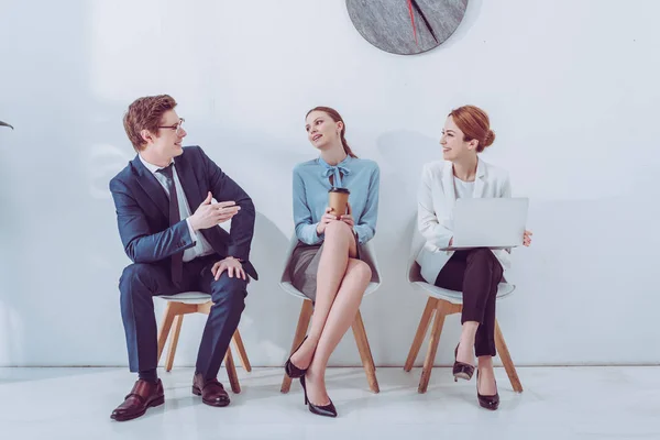 Hombre alegre en gafas hablando con mujeres atractivas esperando entrevista de trabajo - foto de stock